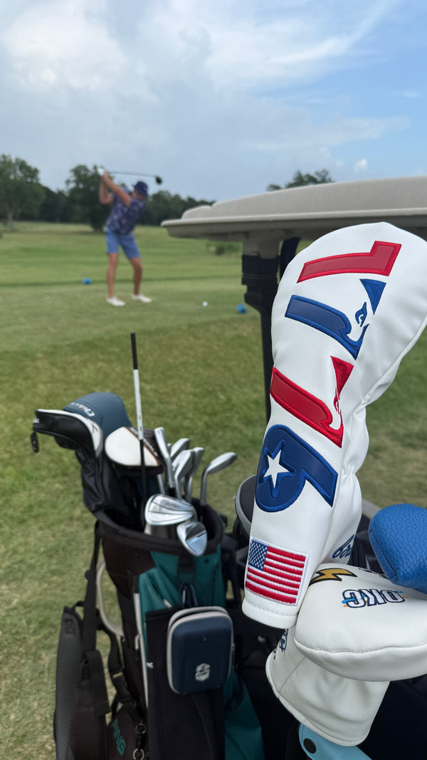 A white golf club headcover with red and blue embroidered graphics, including a stylized 'F' logo and an American flag patch near the bottom.